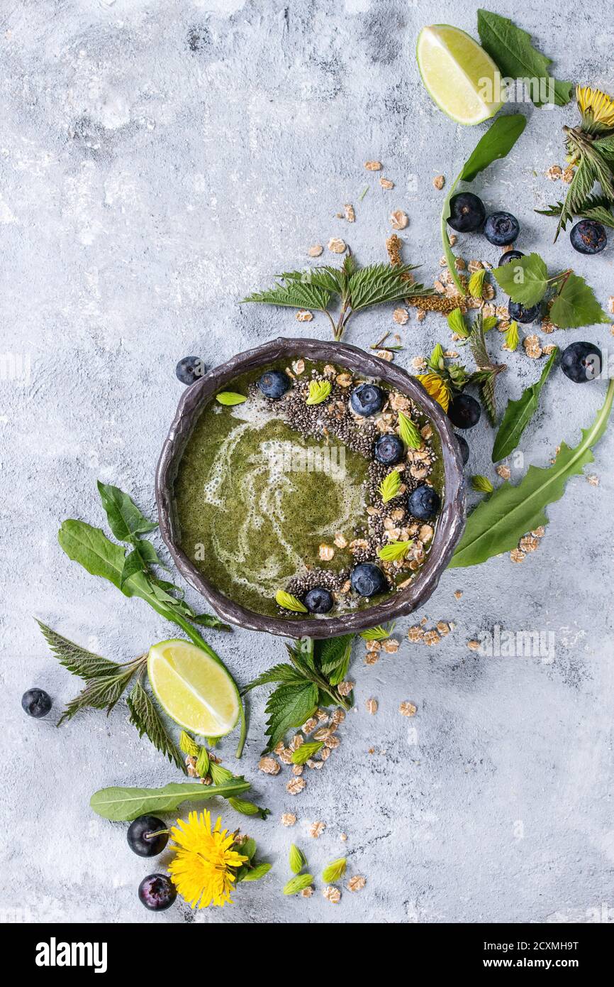 Verde primavera ortica e tarassaco ciotola smoothie servita con calce, fiori gialli, foglie giovani, fiocchi di avena, chia semi, mirtilli, crema su grigio b Foto Stock