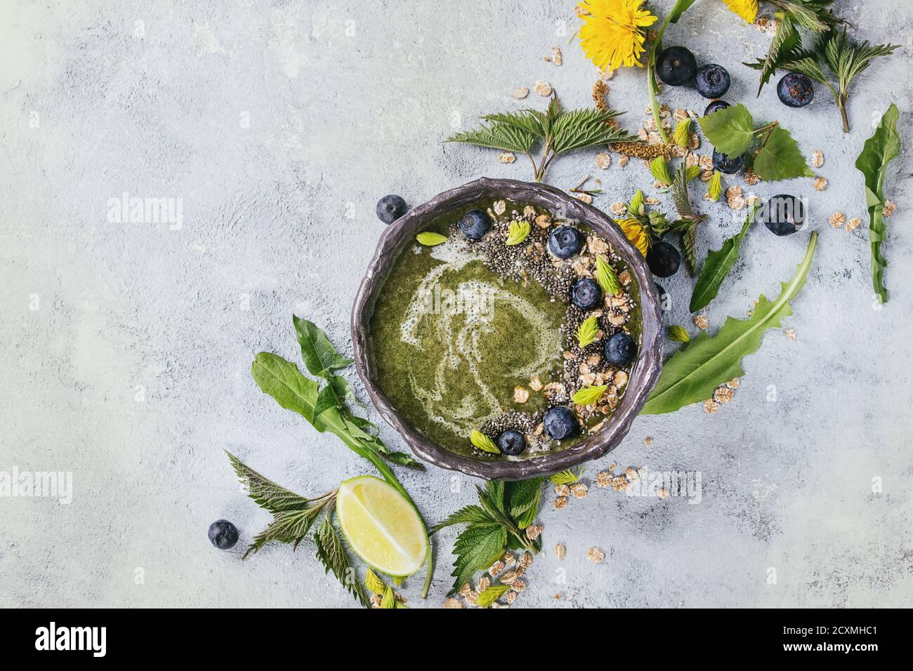 Verde primavera ortica e tarassaco ciotola smoothie servita con calce, fiori gialli, foglie giovani, fiocchi di avena, chia semi, mirtilli, crema su grigio b Foto Stock