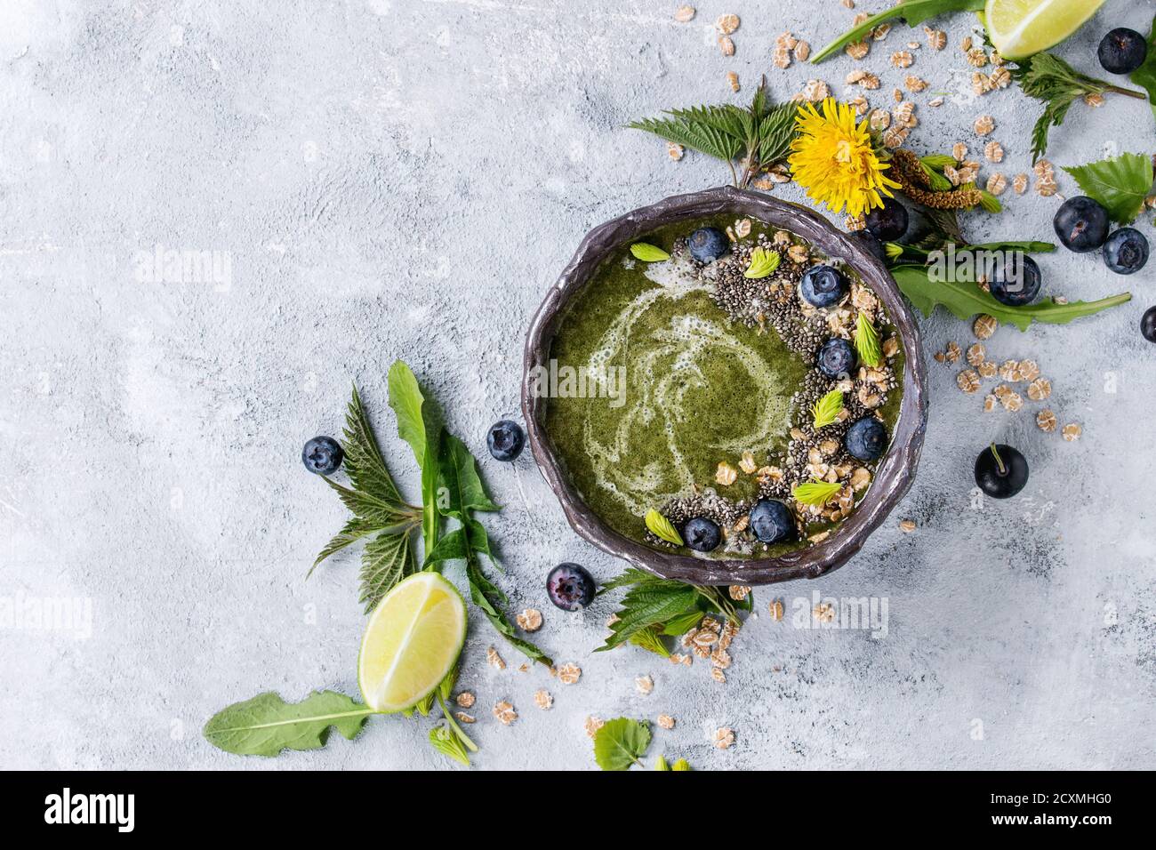 Verde primavera ortica e tarassaco ciotola smoothie servita con calce, fiori gialli, foglie giovani, fiocchi di avena, chia semi, mirtilli, crema su grigio b Foto Stock