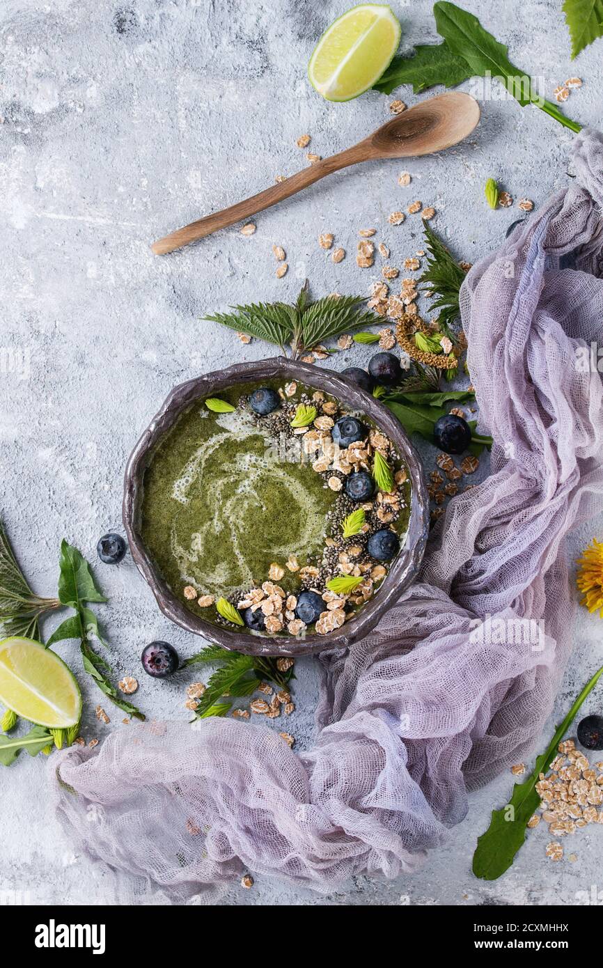 Verde primavera ortica e tarassaco ciotola smoothie servita con calce, fiori gialli, foglie giovani, fiocchi di avena, semi di Chia, mirtilli, garza e cucchiaio o Foto Stock