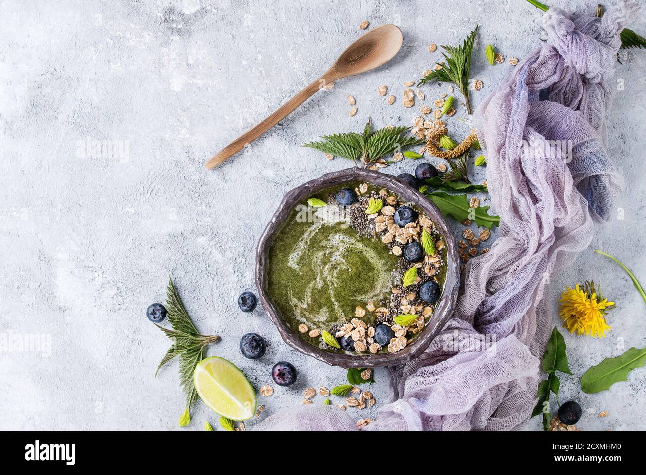 Verde primavera ortica e tarassaco ciotola smoothie servita con calce, fiori gialli, foglie giovani, fiocchi di avena, semi di Chia, mirtilli, garza e cucchiaio o Foto Stock
