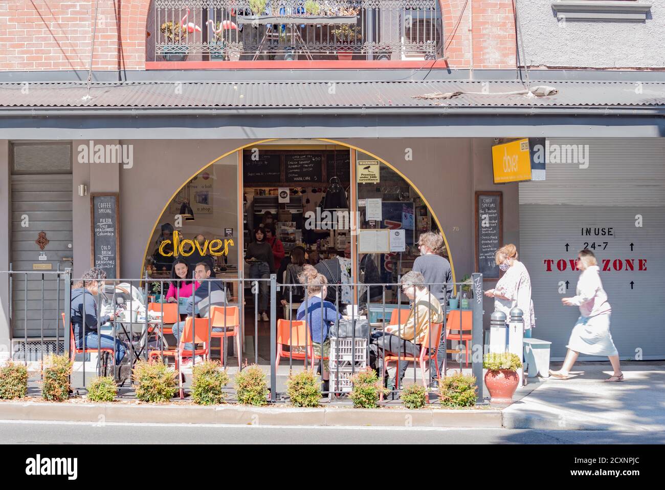 La gente siede socialmente distanziata fuori di un caffè mentre una donna che indossa una maschera cammina attraverso il sobborgo di Sydney di Annandale, nuovo Galles del Sud, Australia Foto Stock