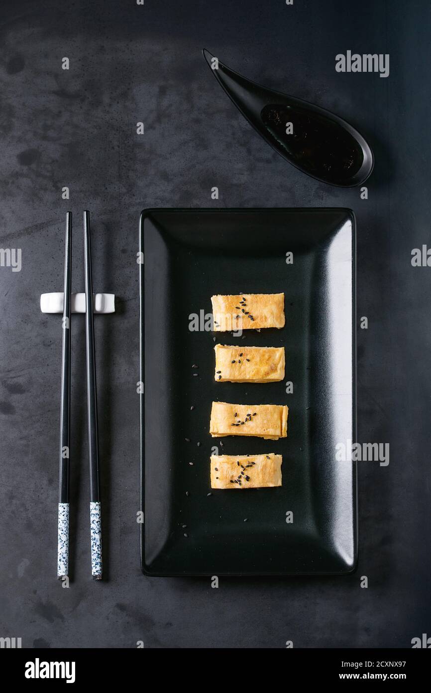 Giapponese frittata arrotolata Tamagoyaki affettata con seasame nero semi e la salsa di soia, servita nel quadrato nero piastra in ceramica con bacchette su metallo scuro Foto Stock