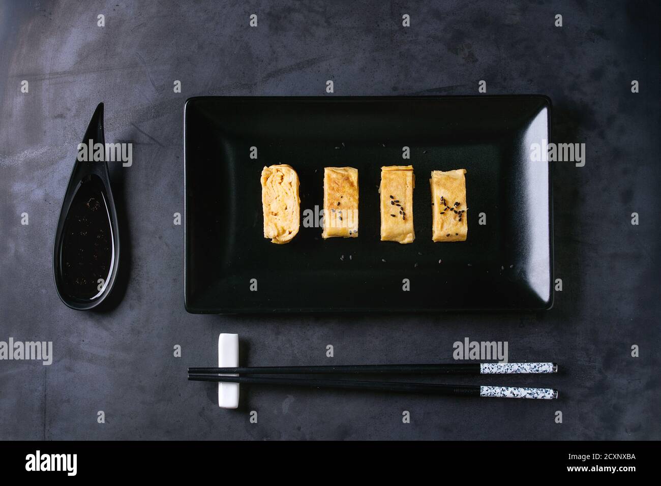 Giapponese frittata arrotolata Tamagoyaki affettata con seasame nero semi e la salsa di soia, servita nel quadrato nero piastra in ceramica con bacchette oltre il buio meta Foto Stock