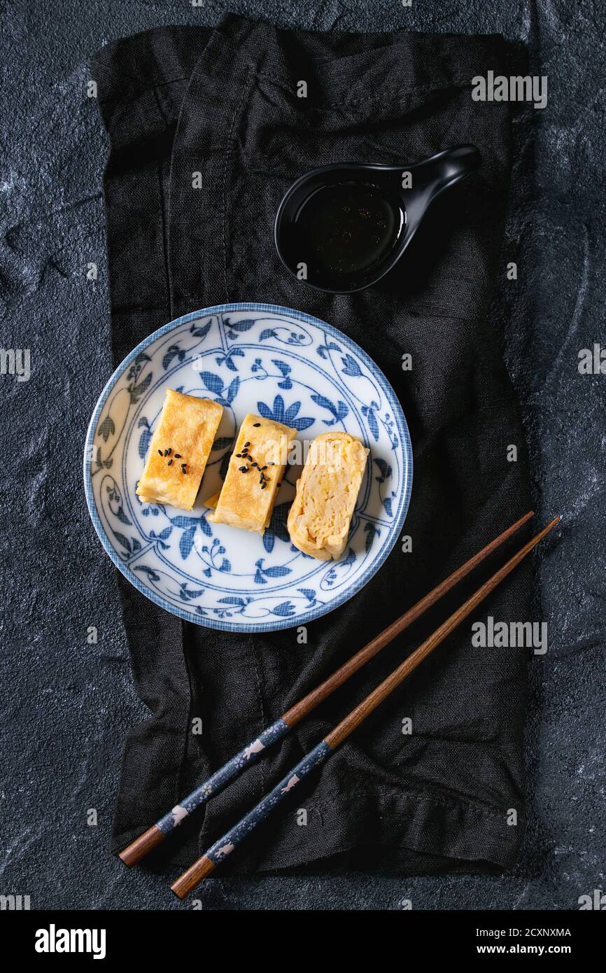 Giapponese laminato Omelet Tamagoyaki affettato con semi di condimenti nero e salsa di soia, servito in bianco blu piatto di ceramica ornata con chopsticks, tessuto ov Foto Stock