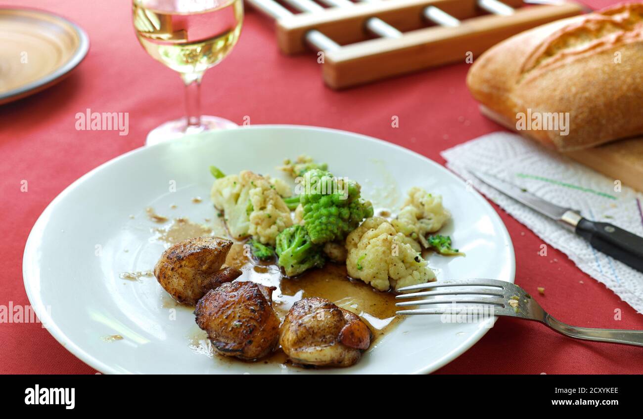 Delizioso piatto francese: capesante coquilles St Jacques con verdure al vapore su un piatto bianco, pane e un bicchiere di vino bianco sul tavolo rosso Foto Stock