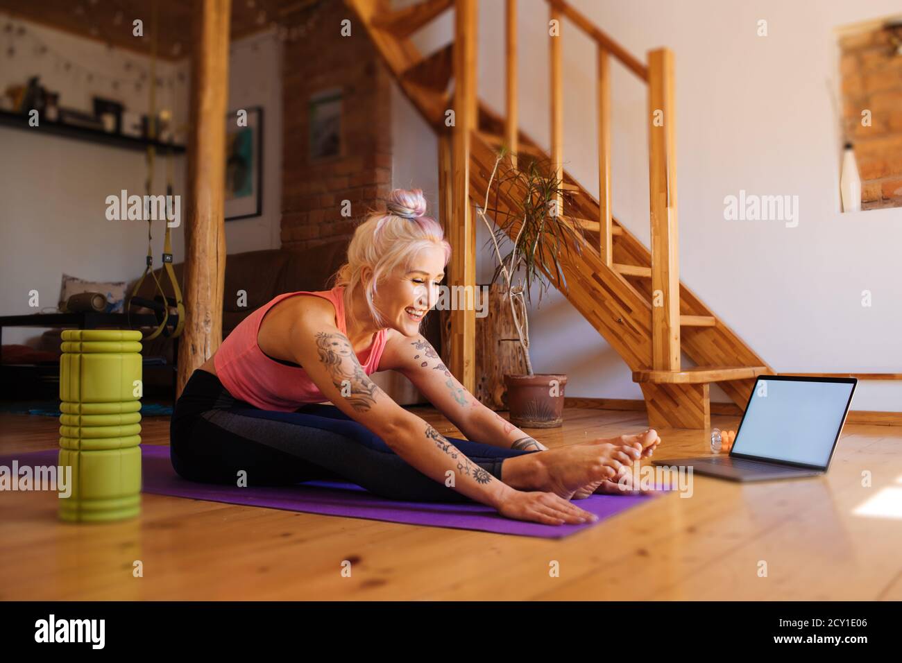 Giovane donna con un tatuaggio e rullo in schiuma da parte. Fitness esercizio virtuale online. Stretching da internet a casa. Una ragazza sportiva in abbigliamento sportivo Foto Stock