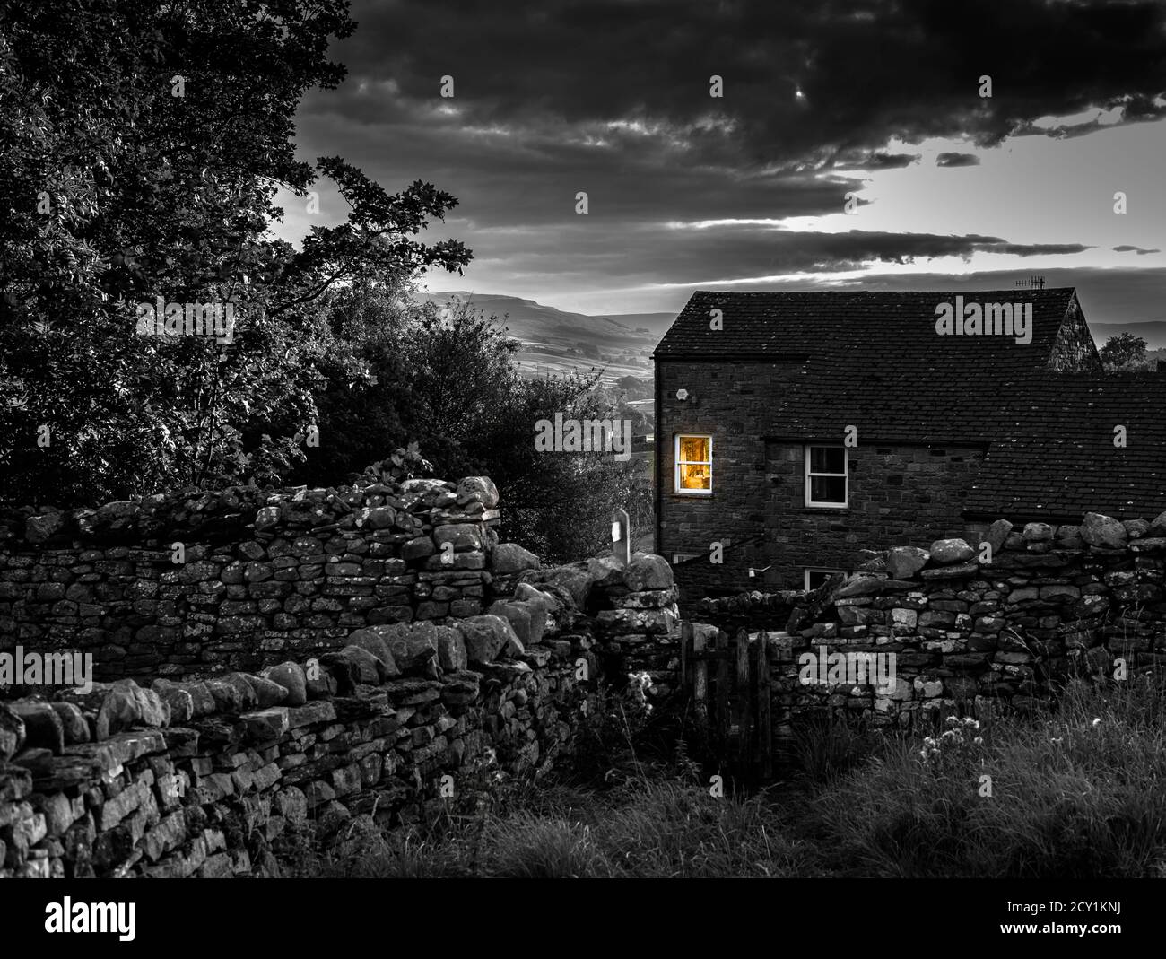 Immagine in bianco e nero di un cottage in pietra al tramonto con un caldo bagliore arancione da una finestra nelle Yorkshire Dales, Inghilterra, Regno Unito. Foto Stock
