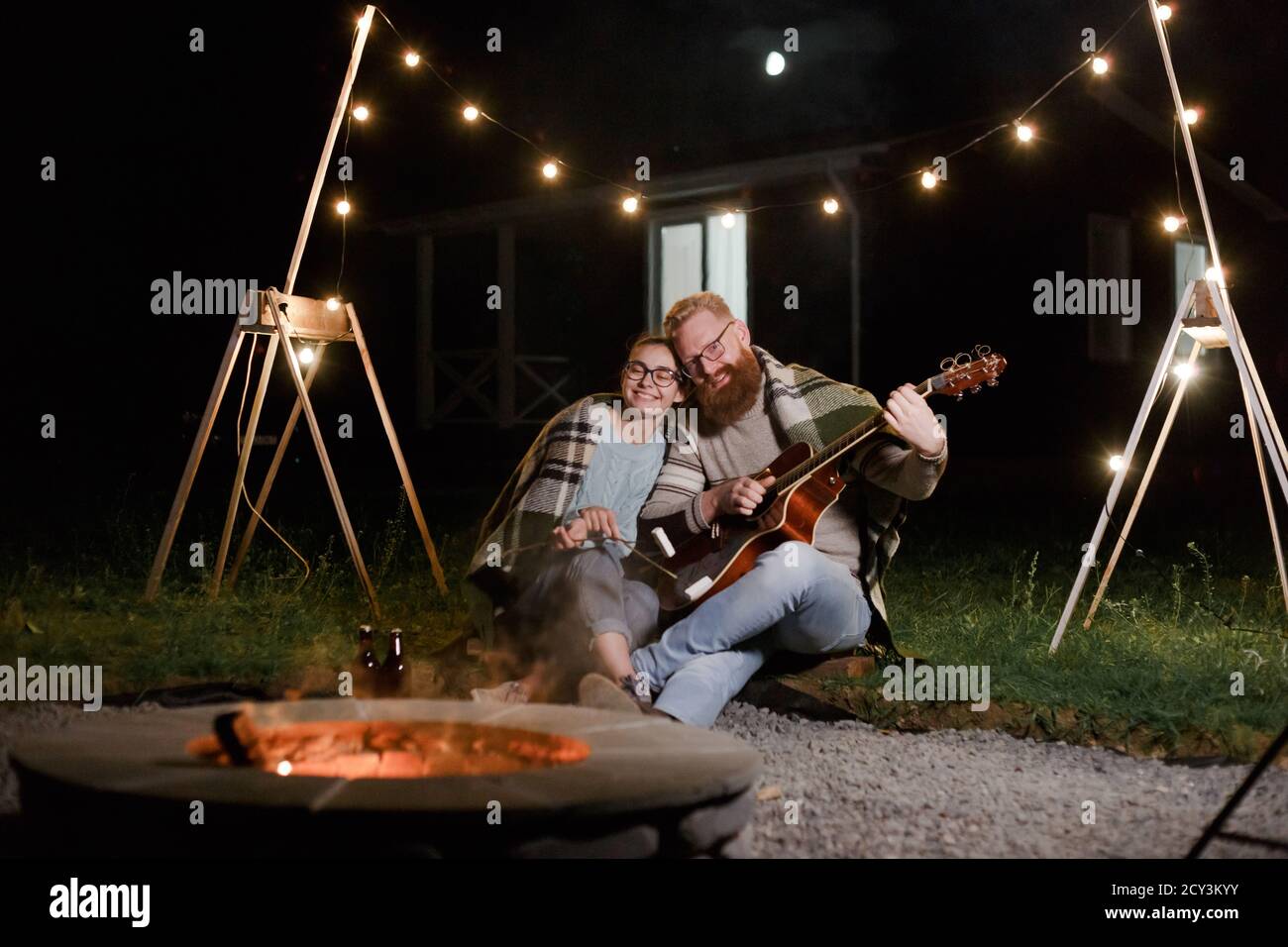 Giovane coppia, donna caucasica e uomo bearded dai capelli rossi, in un pic-nic notturno con una chitarra e marshmello hanno un buon tempo Foto Stock