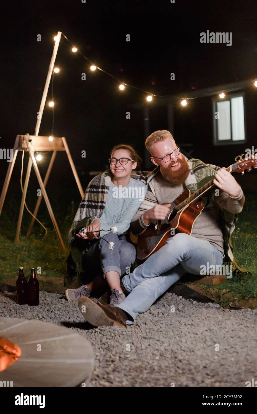 Giovane coppia, donna caucasica e uomo bearded dai capelli rossi, in un pic-nic notturno con una chitarra e marshmello hanno un buon tempo Foto Stock