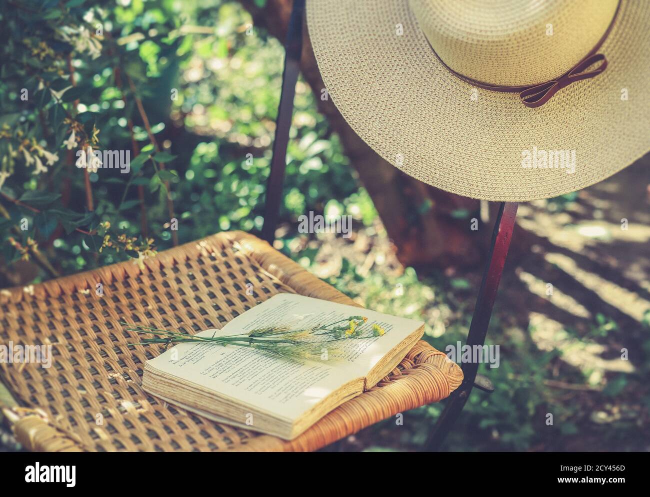 Fiori di campo su libro aperto e cappello estivo femminile su vecchia sedia in vimini in giardino, filtre retrò, concetto estetico nucleo cottage Foto Stock