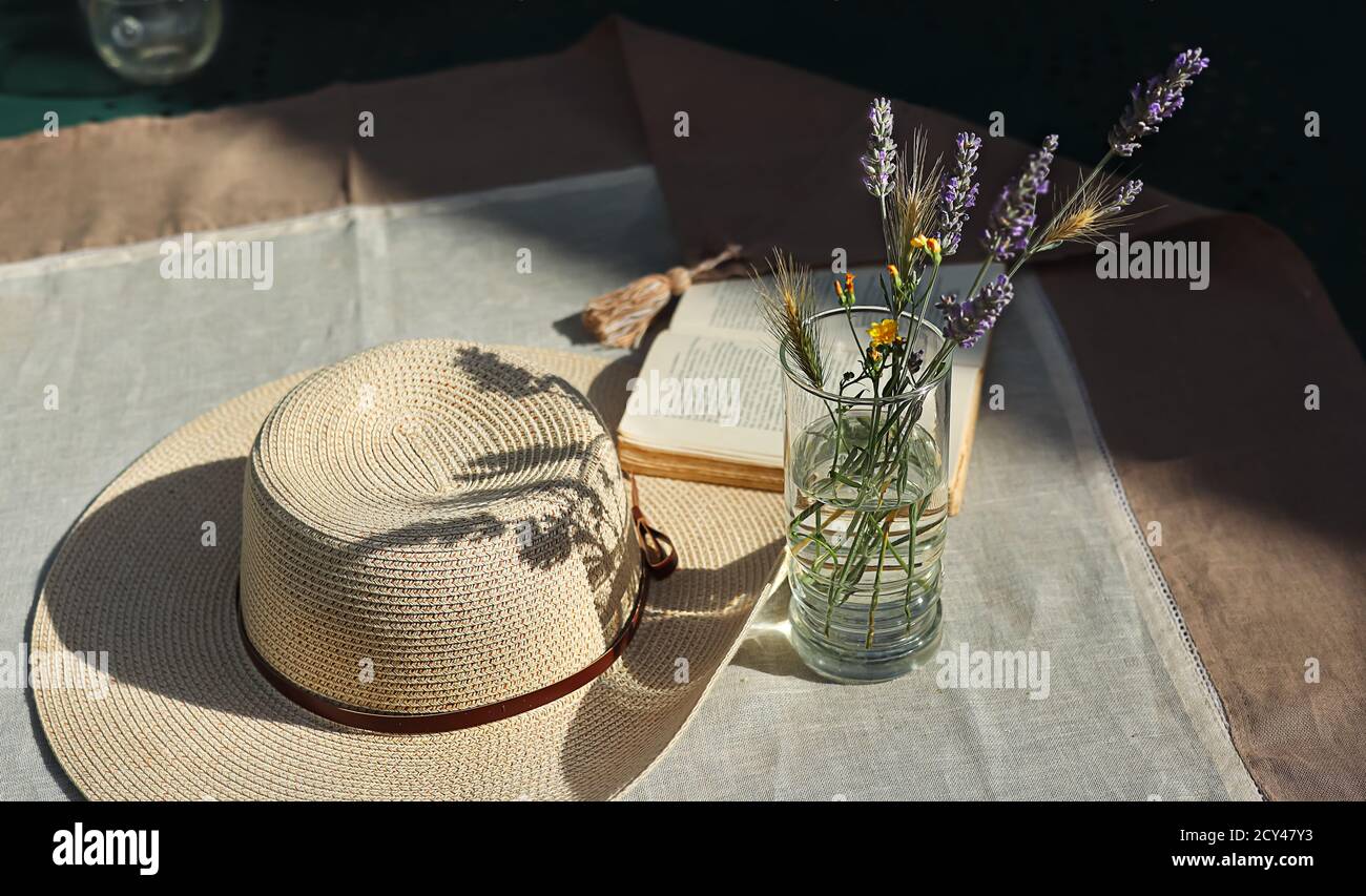 Fiori selvatici nel bicchiere con acqua, un libro aperto e cappello estivo femminile con ribone su un tavolo coperto di tovaglia di lino con le ombre della su Foto Stock