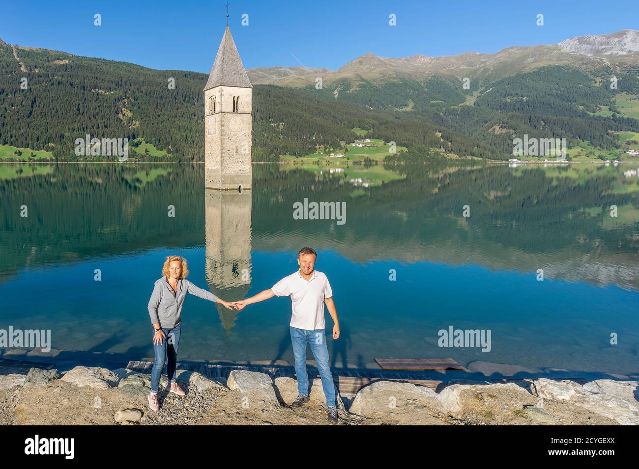 Un paio di turisti si posano di fronte al vecchio campanile di Curon Venosta, che si riflette perfettamente nelle acque fertose del Lago di Resia, Alto Adige, Foto Stock