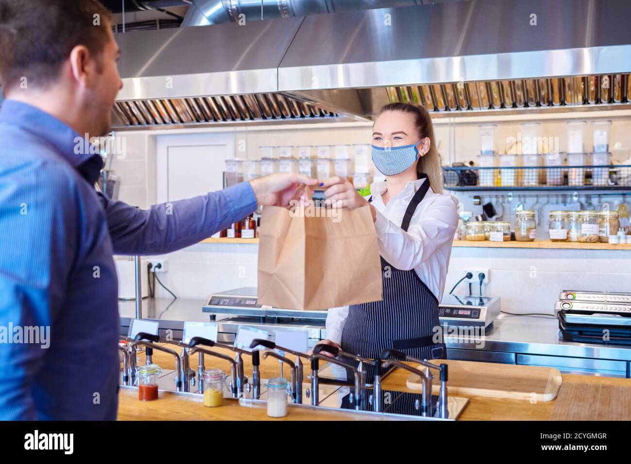Cameriera felice che indossa una maschera protettiva che serve cibo da asporto al cliente al banco del piccolo ristorante per famiglie. Foto Stock