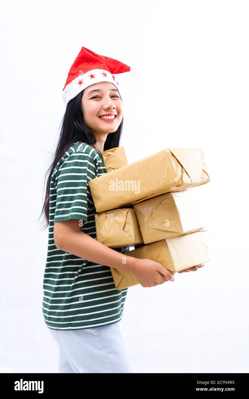 Ritratto di una giovane donna asiatica che indossa un cappello di santa e indossare abiti casual che sollevano un mucchio di regali con uno sfondo bianco isolato Foto Stock