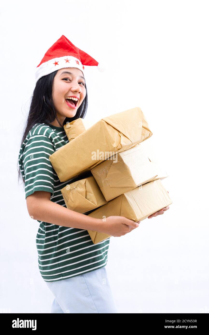 Ritratto di una giovane donna asiatica che indossa un cappello di santa e indossare abiti casual che sollevano un mucchio di regali con faccia a sorpresa in uno sfondo bianco isolato Foto Stock