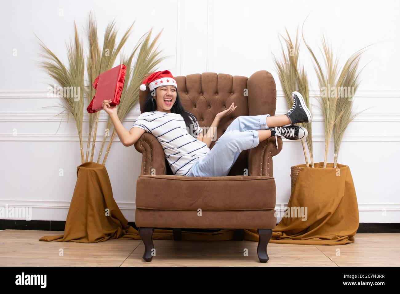 Ritratto di giovane donna asiatica che indossa un cappello di Santa e. indossando abiti informali seduti su un divano marrone all'interno Foto Stock