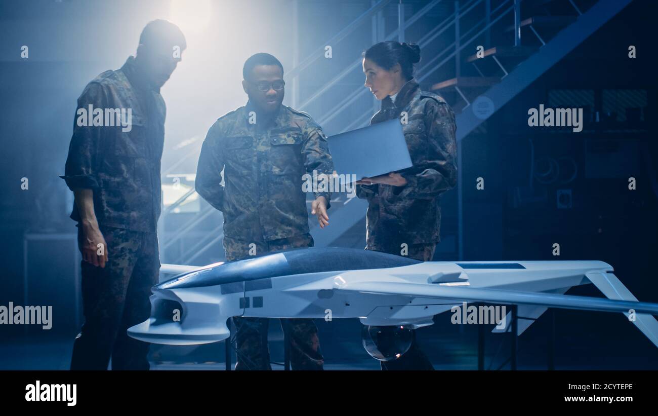 Gli ingegneri aerospaziali dell'esercito lavorano al drone di un veicolo aereo senza equipaggio. Esperti di aviazione in uniforme parlano, usando il laptop. Impianto industriale con aeromobili per Foto Stock