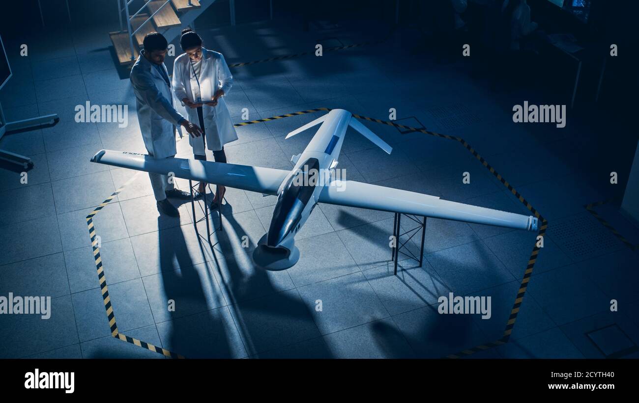 Due ingegneri aerospaziali lavorano sul prototipo di drone di un veicolo aereo senza equipaggio. Scienziati dell'aviazione in White Coats che parlano, utilizzando Tablet computer. Industriale Foto Stock