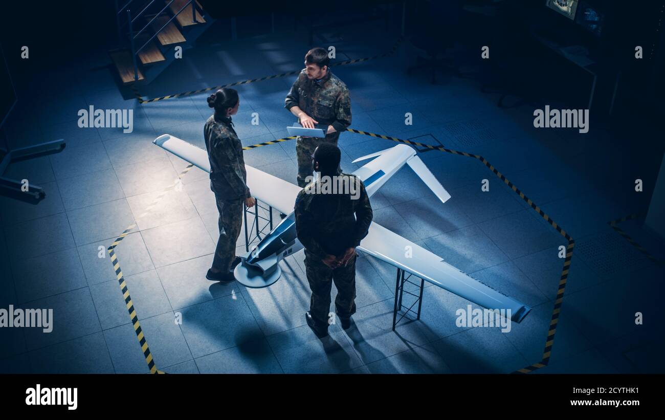 Gli ingegneri aerospaziali dell'esercito lavorano al drone di un veicolo aereo senza equipaggio. Esperti di aviazione in uniforme parlano, usando il laptop. Struttura industriale con sorveglianza Foto Stock