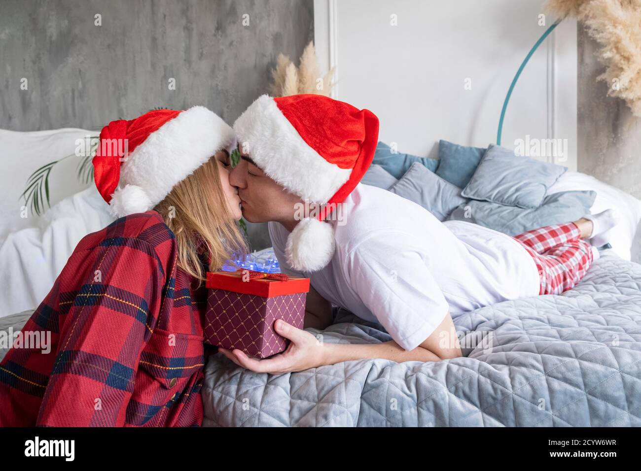 Vacanze invernali e concetto di persone. Felice coppia caucasica amorevole con regalo di natale baciare a casa sdraiata a letto indossando cappelli santa Foto Stock