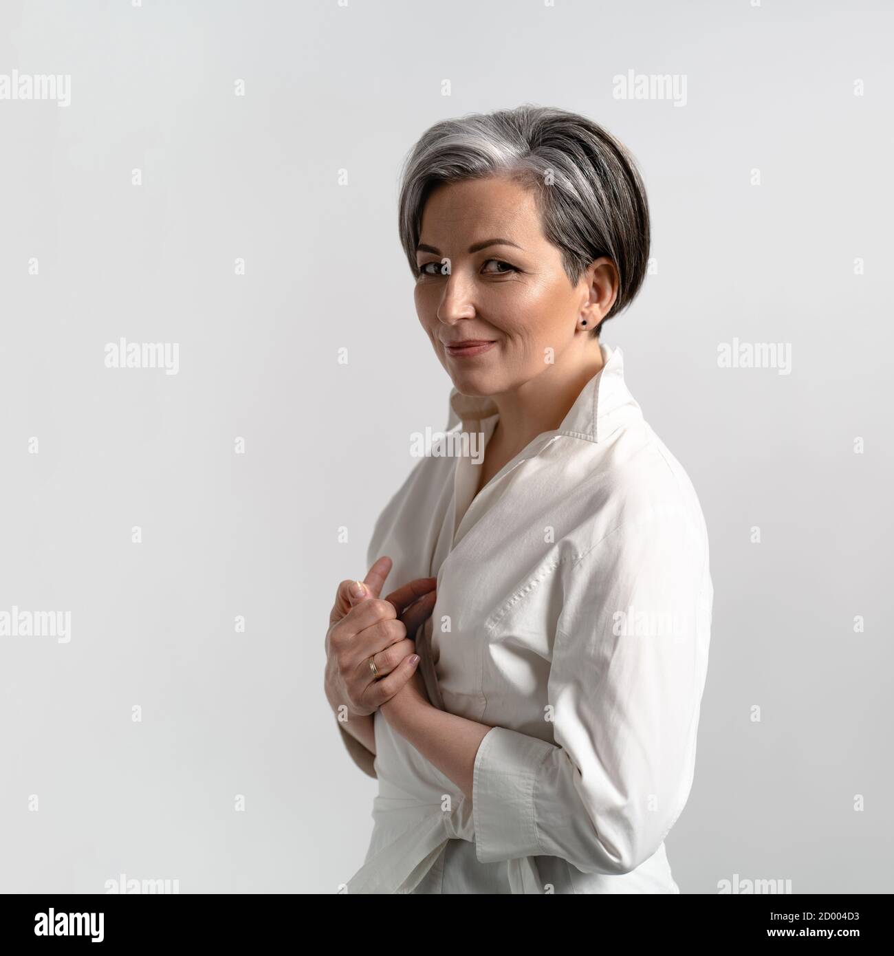 Donna di bellezza in camicia bianca su uno sfondo isolato in studio. Donna con capelli grigi ha piegato le braccia e sorride alla macchina fotografica. Foto di alta qualità Foto Stock