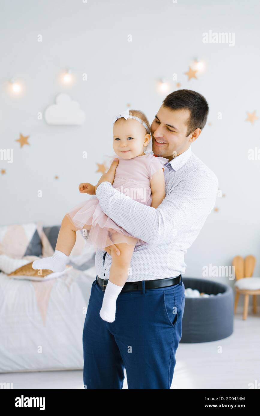 Il giovane papà felice tiene una figlia sorridente di un anno nella sua braccia nella stanza dei bambini Foto Stock