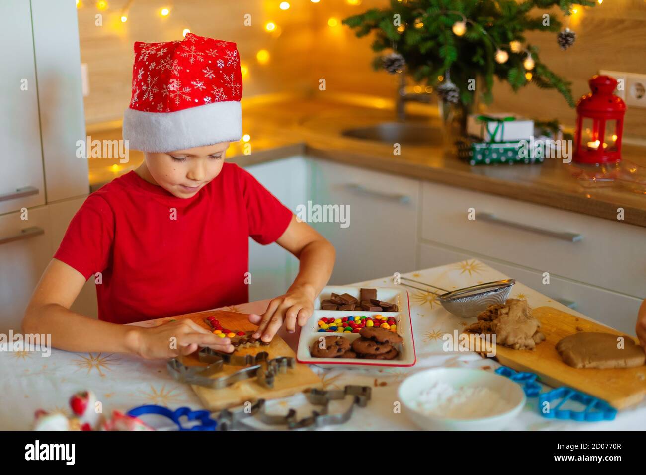 Un ragazzo in un cappello babbo natale prepara i biscotti in cucina. Forma per pan di zenzero di Natale. Bambino in cucina nel decor di Capodanno. Biscotti di Natale Foto Stock