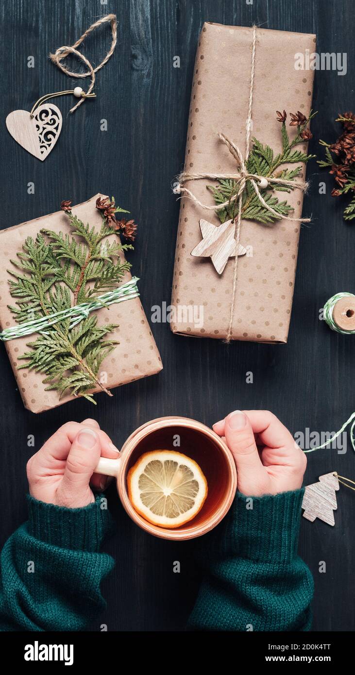 Scatole regalo di Natale avvolte in carta kraft e decorare con rami di cipresso su rustico sfondo di legno. Tazza da donna con tè. Vista dall'alto Foto Stock