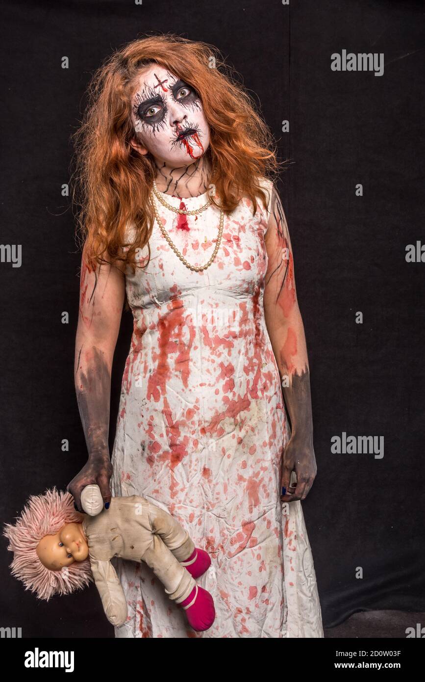 Una terrificante testa rossa in piedi con una bambola in mano, che indossa abiti bianchi sanguinosi. Concetto di Halloween Foto Stock