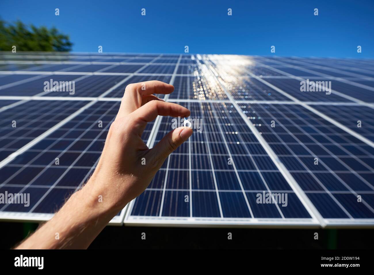 FrontView della mano del lavoratore che tiene minuscolo dettaglio per l'installazione della batteria solare di fronte al pannello solare su sfondo. Tendine parasole su superficie liscia. Esterni high-tech innovativi. Ambiente amichevole. Foto Stock
