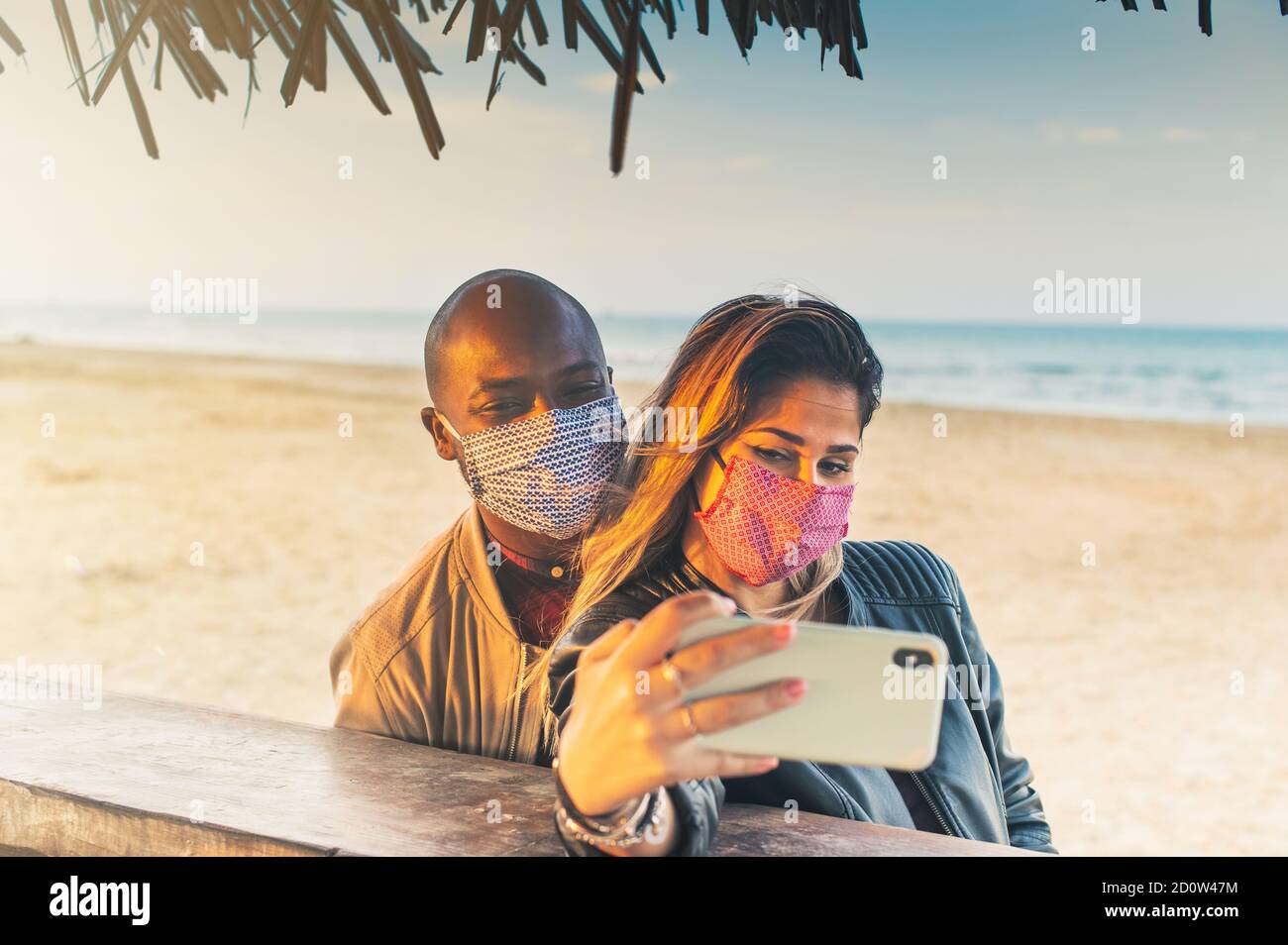 Amici millenari multirazziali che prendono selfie sorridendo dietro le maschere di faccia a. Bar sulla spiaggia - amicizia felice e nuovo normale concetto con i giovani Foto Stock