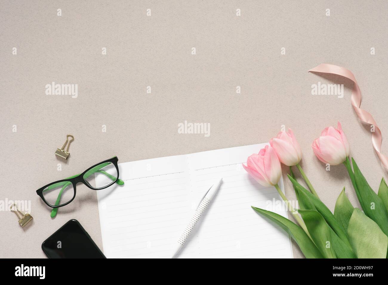 Home Office Desk in tonalità rosa polverosa con telefono, tulipani rosa, bicchieri, pagine pulite di un diario aperto e penne su uno sfondo di caffè. Lavori di posa piatta f Foto Stock