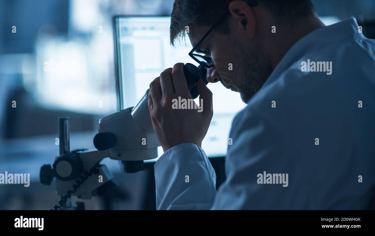 Shot of a Male IT Scientist utilizza un computer con microscopio elettronico. Nel background Technology Development Laboratory con scienziati, ingegneri Foto Stock