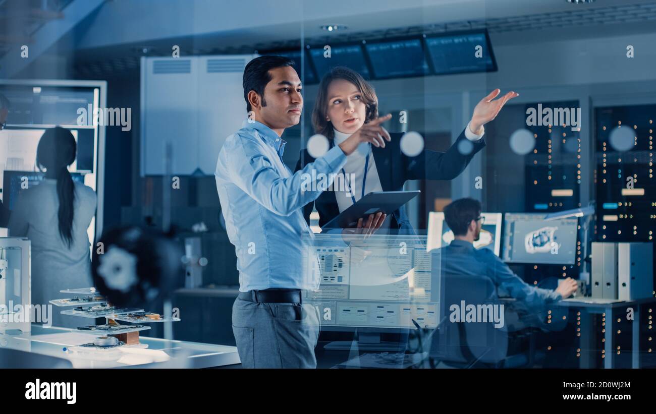 In Technology Research Facility: Il Project Manager femminile parla con il Chief Engineer, consulta il Tablet computer. Team di ingegneri industriali Foto Stock