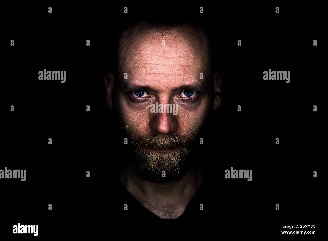 Un uomo guarda seriamente nella macchina fotografica dall'oscurità. Il suo sguardo è minaccioso e determinato. Foto Stock
