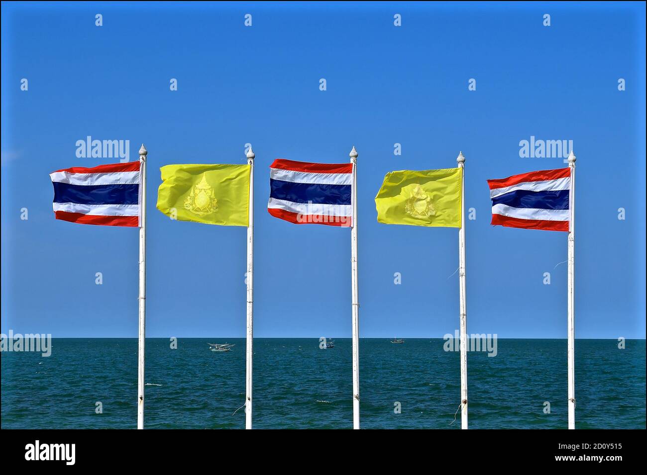 Una fila di bandiere thailandesi e la bandiera gialla del re che battono in un forte vento onshore contro uno sfondo di cielo blu e mare. Foto Stock