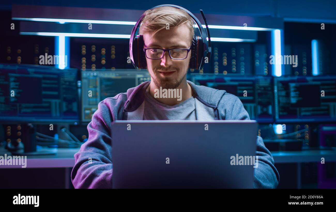 Ritratto dello sviluppatore di software Hacker Gamer indossando occhiali e cuffie seduti alla sua scrivania e lavorando giocando sul laptop. In background scuro Alto Foto Stock
