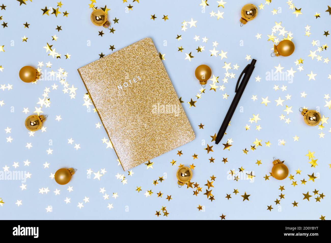 Taccuino d'oro con copertina lucida glitter, penna nera e palline d'oro natalizie, baubles e stelle confetti su sfondo blu. Lista dei desideri, Capodanno reso Foto Stock