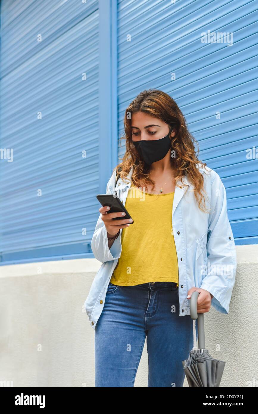 ritratto verticale di una giovane donna rossa che indossa un nero maschera in piedi in strada con ombrello in una mano e. controllare il suo smartphone vicino a un blu Foto Stock