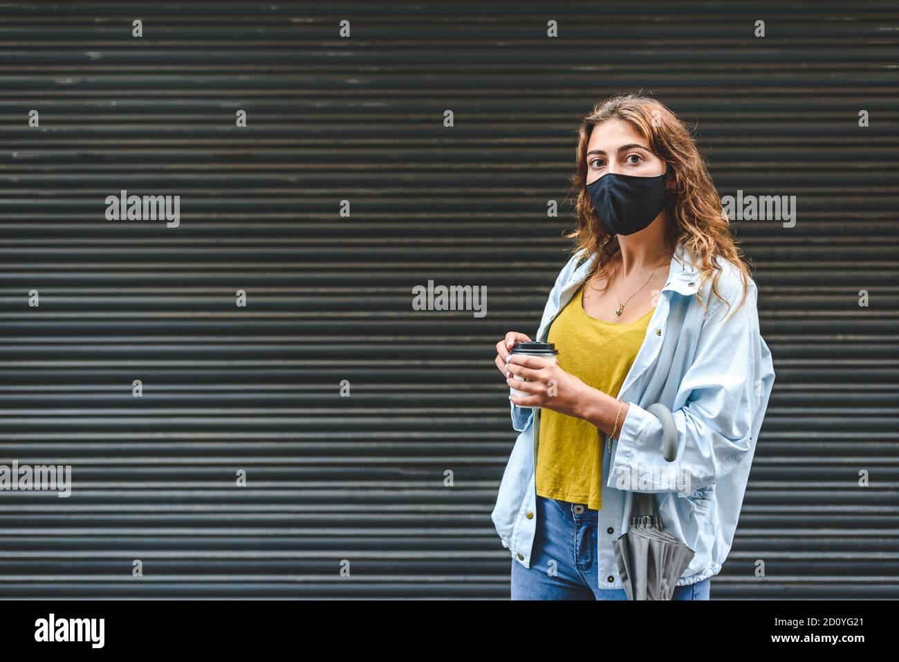 giovane donna caucasica rossa con una maschera nera che rimane sulla strada con una tazza di carta di take away caffè con sfondo nero Foto Stock