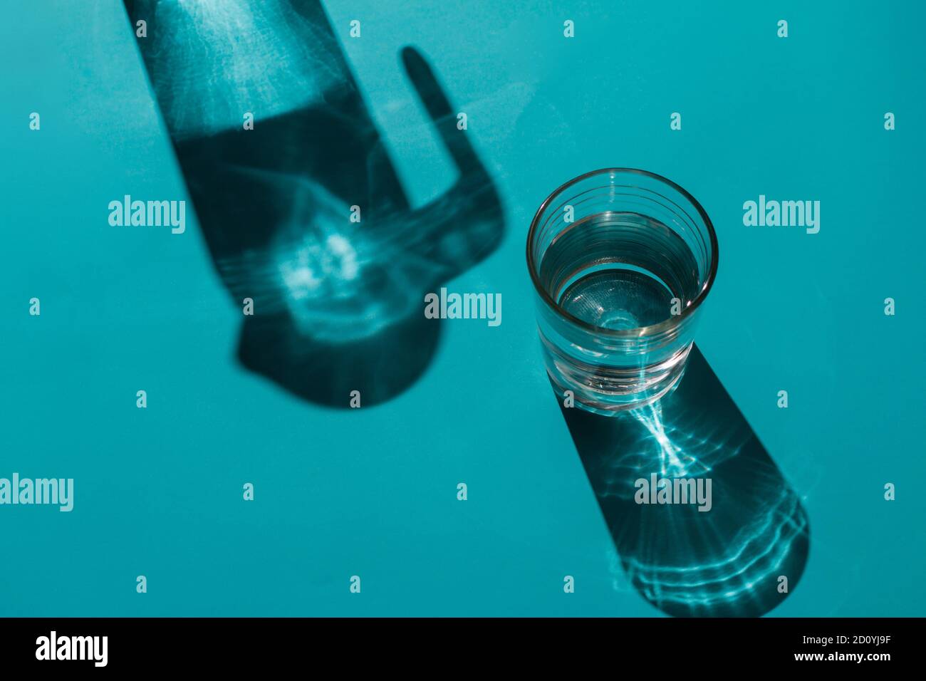 Un bicchiere con acqua accanto a una caraffa su un sfondo blu chiaro in cui vengono proiettate le ombre Foto Stock