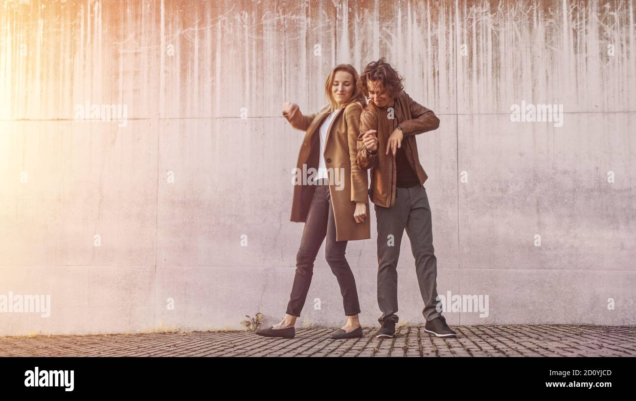 Cheerful Girl e Happy Young Man with Long Hair stanno ballando attivamente su una strada accanto a un muro urbano di cemento. Indossano giacca in pelle marrone e. Foto Stock