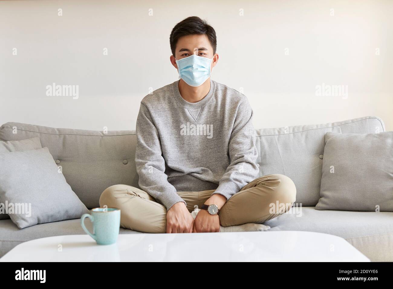 giovane uomo asiatico che rimane a casa indossando la maschera seduta sopra la gamba del lettino ha attraversato la telecamera Foto Stock