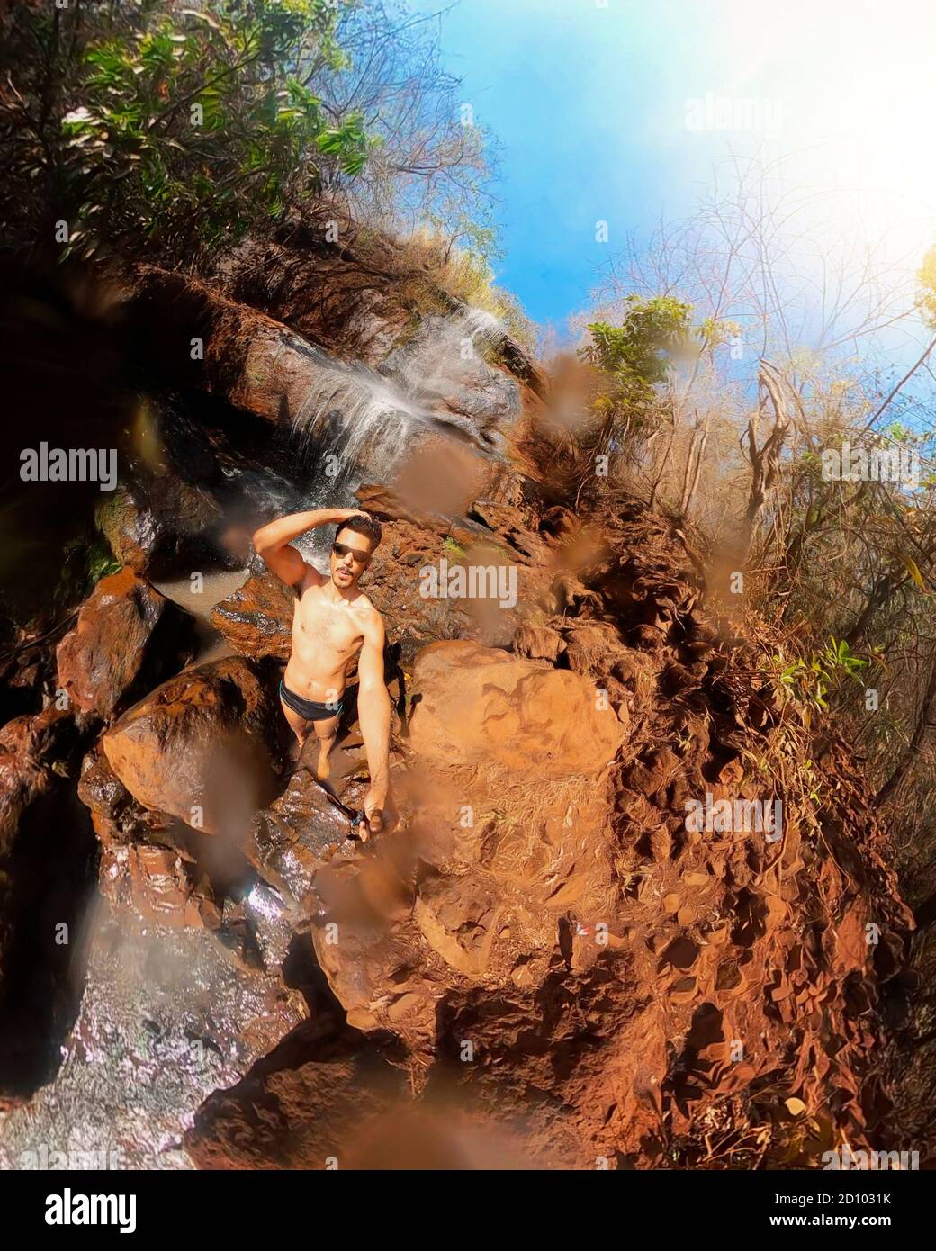 Uomo in occhiali e bunks nuoto alla cascata con 360 fotocamera in una giornata di sole Foto Stock