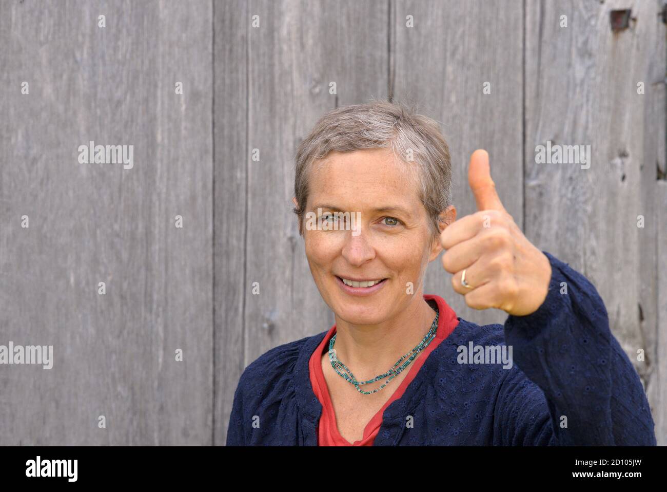 Sorridente contadina caucasica di mezza età mostra un grumo (segno di approvazione) con emozioni ambigue, di fronte ad un vecchio fienile di legno di fondo. Foto Stock