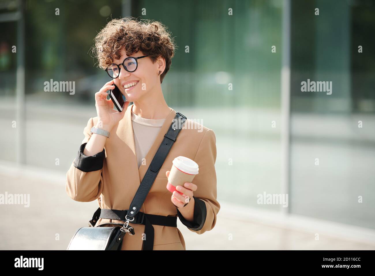 Giovane donna d'affari allegra con drink in mano e smartphone da orecchio Foto Stock