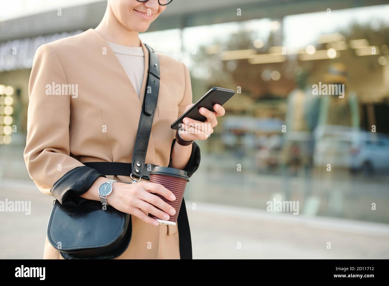 Giovane donna sorridente con bevanda e borsa che scorre attraverso i contatti o sms Foto Stock