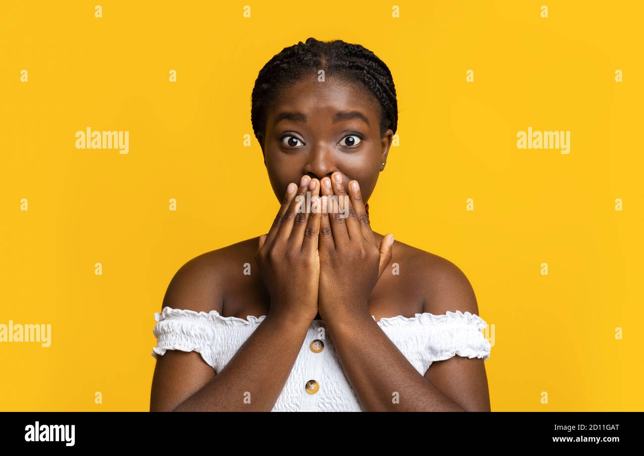 Scioccata African American femmina che ricopre la bocca con le mani, in posa su sfondo giallo Foto Stock