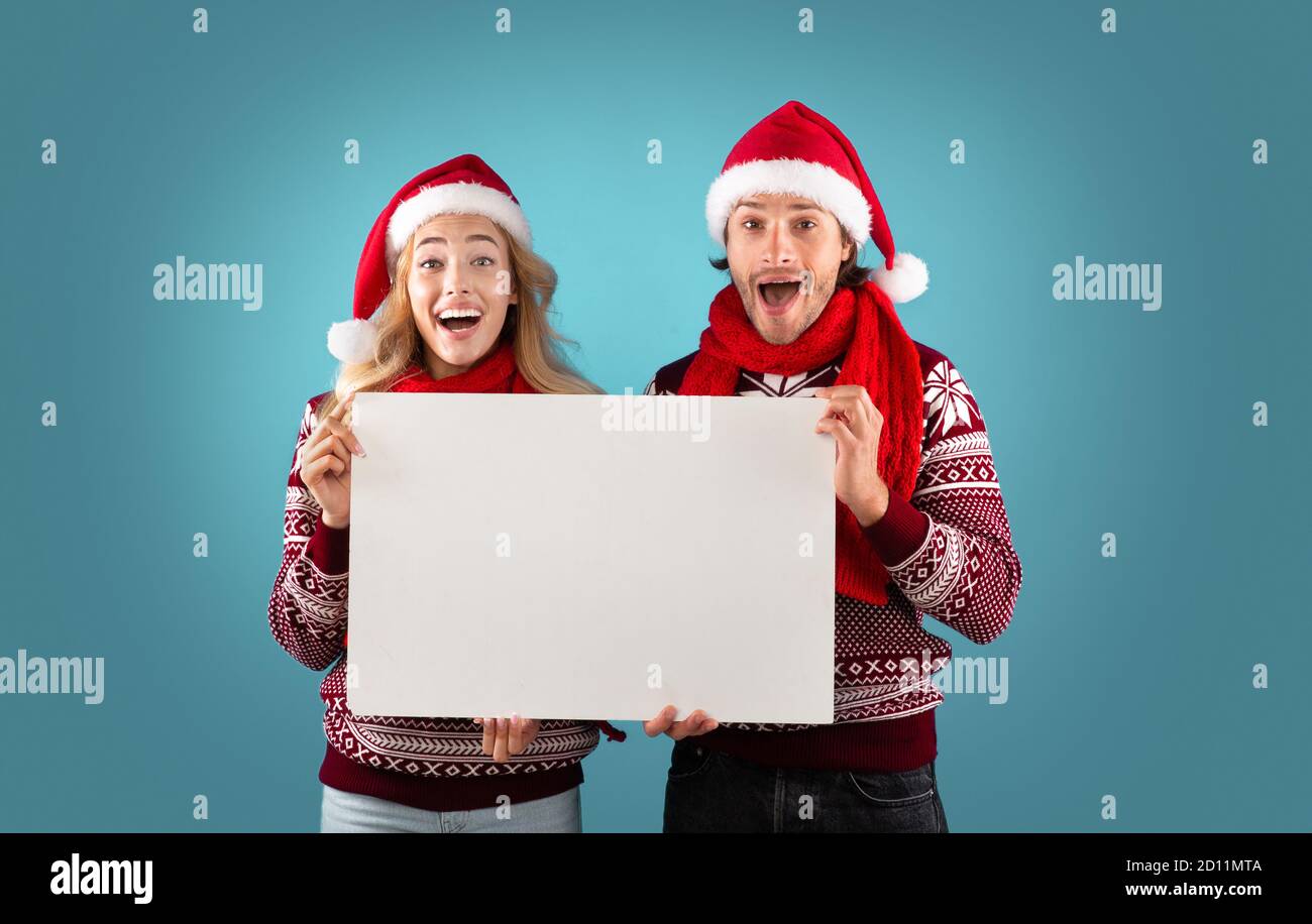 Annuncio di Natale. Coppia millennial entusiasta che tiene un poster vuoto con mockup per il design su sfondo blu Foto Stock